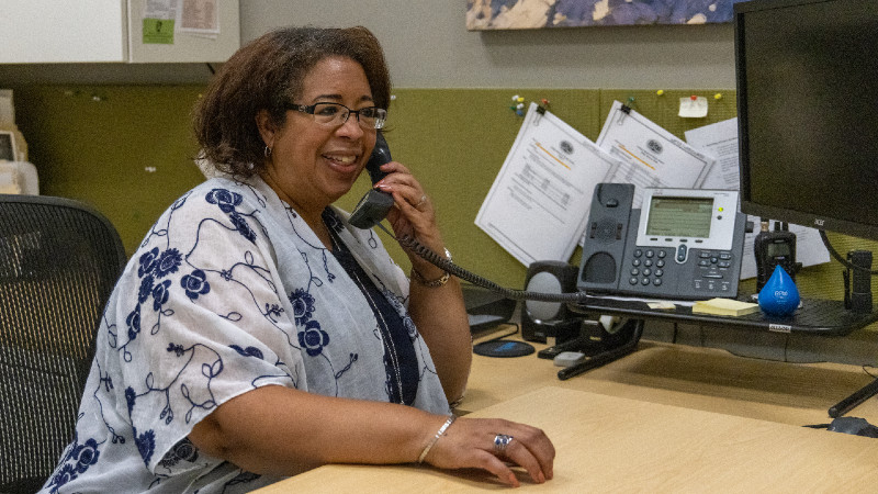 A black woman in a white and blue top answers the phone at her desk.