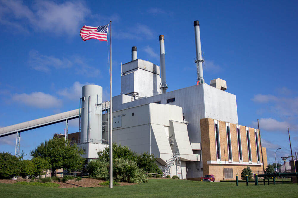 A blocky, steal power plant with a coal escalator and brick facade against a blue sky with an American flag waving in the front.