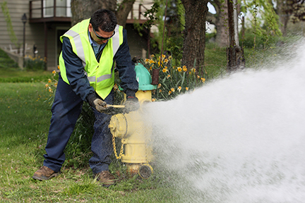 A yellow fire hydrant is spraying water because a man with dark hair who is wearing a yellow reflective vest is performing a water main flush on the hydrant.