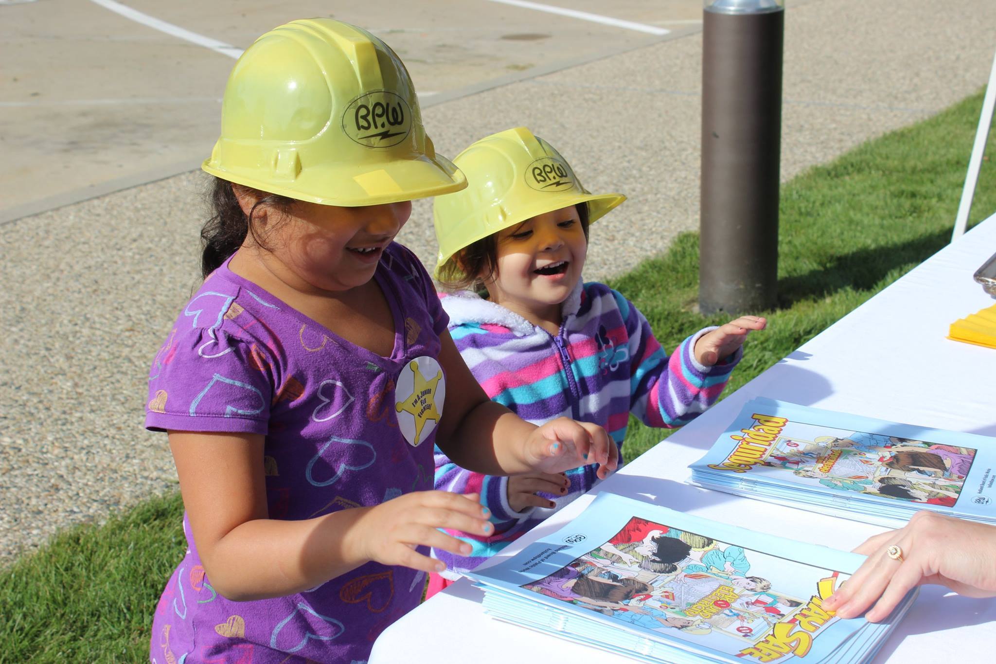 Two little girls smile wearing BPW construction hats. They're standing next to a white table about to pick up a coloring book.