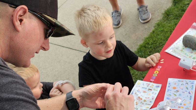 Two little boys create a craft with their dad, who wears a baseball hat and sunglasses.