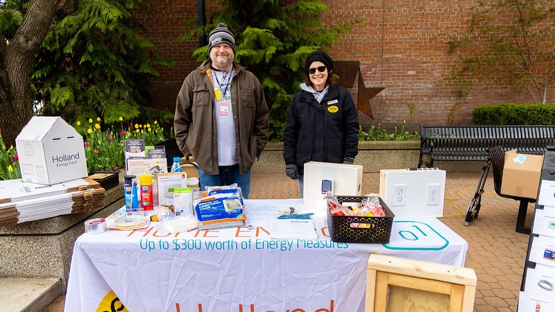 A man and a woman smile behind an outdoor table. They're bundled up and on the table are home efficiency items.