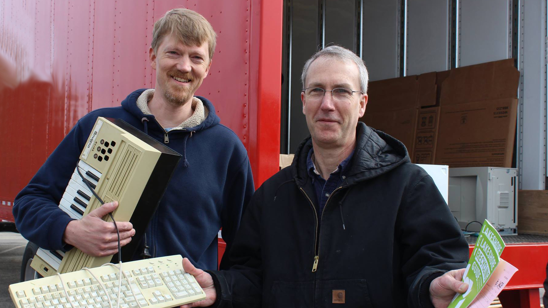 Two men holding old electronics in front of a red truck