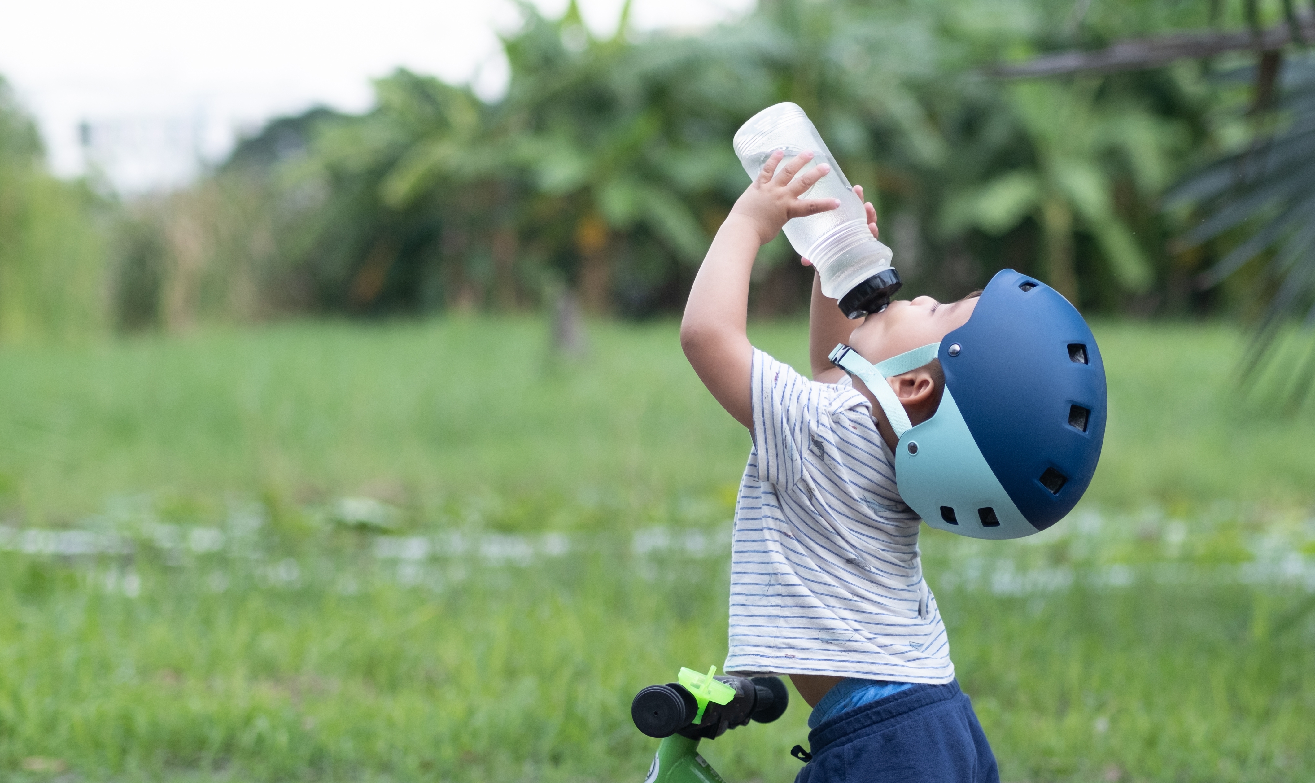 Little boy in a blue bike helmet riding a bike drinks out of a water bottle.