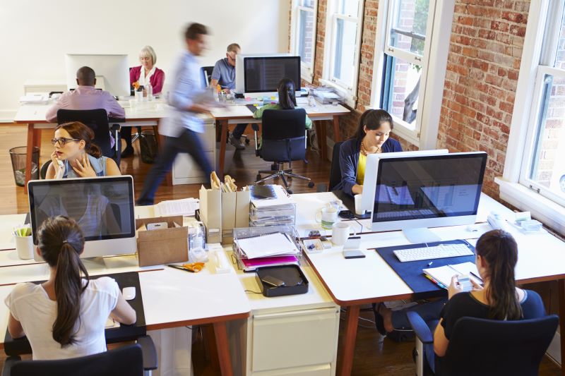 Busy open concept office with people working at desks facing each other. A man streaks past in the background behind them.