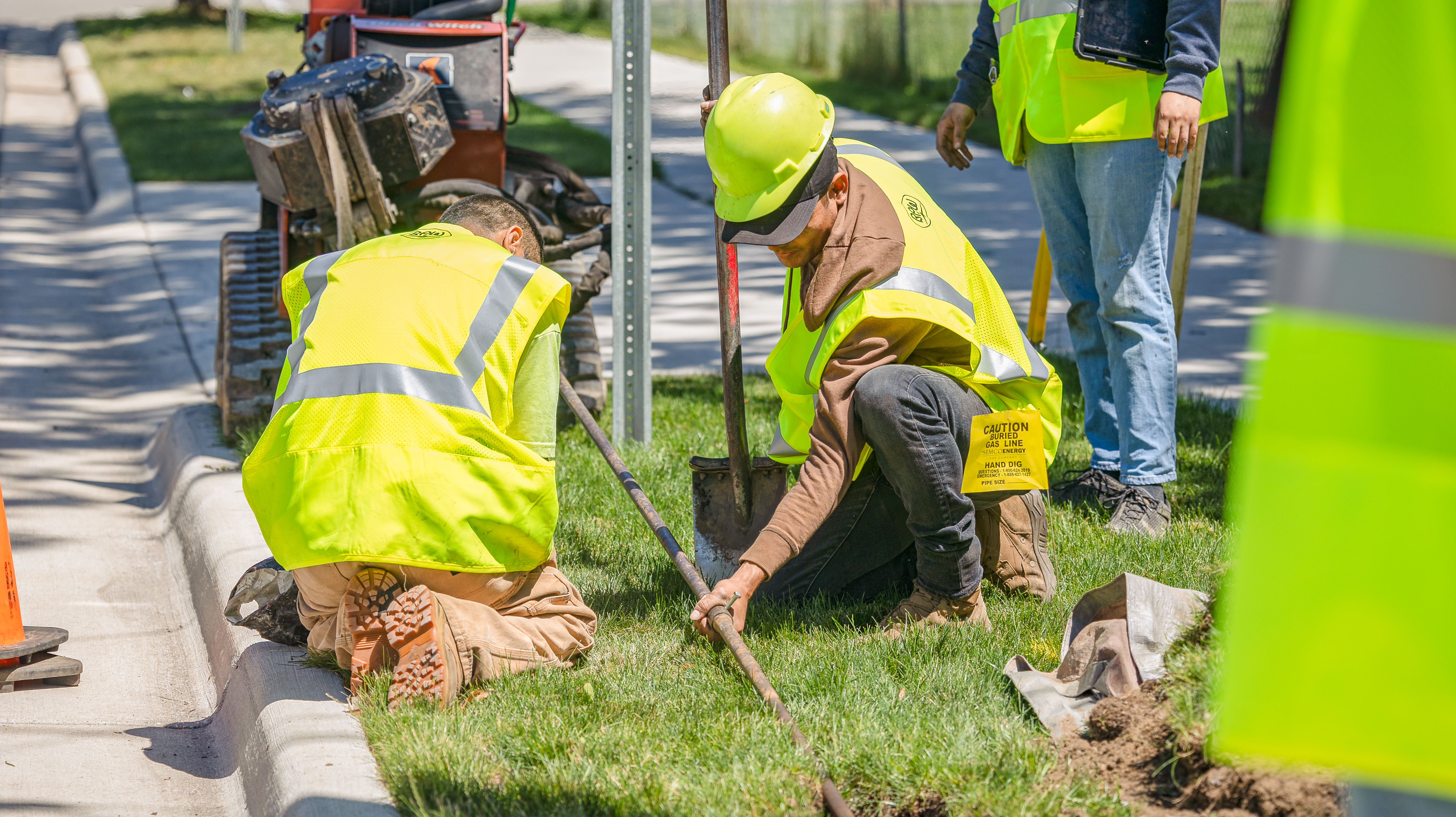 two men wear yellow reflective vests and hard hats while boring a tunnel underground to lay fiber optic cable