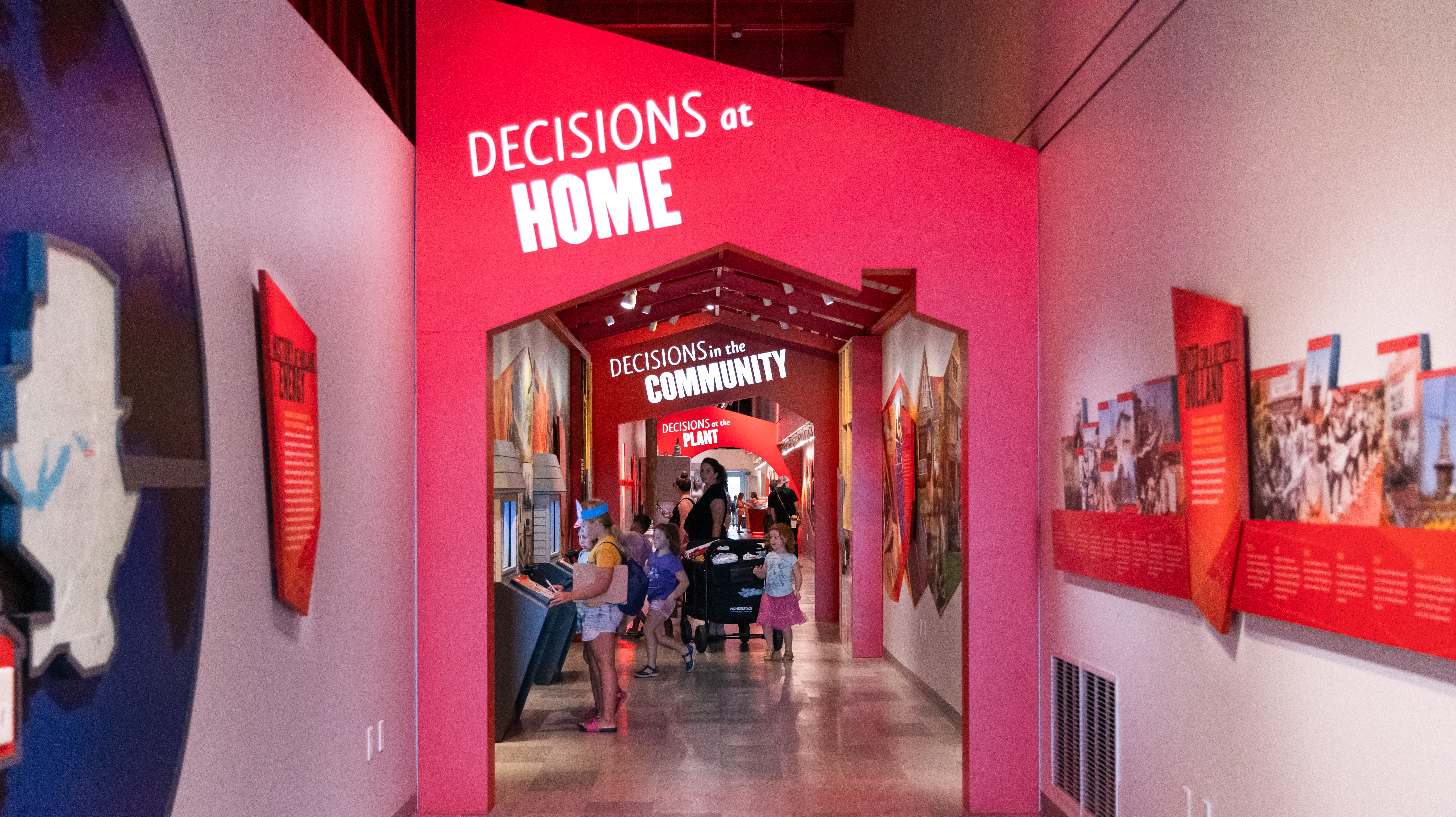 A brightly-lit hallway decorated with warm colored geometric museum displays and a house-shaped kiosk on the left.