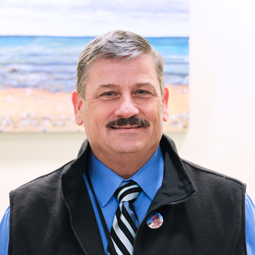 male executive with a moustache wearing a black vest over a blue shirt and striped tie