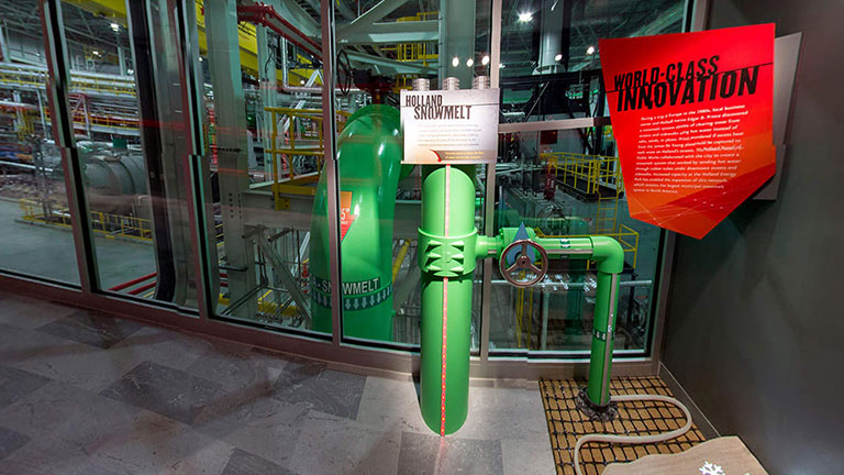 Green snowmelt pipe display in front of a glass window looking into a power plant with a red plaque next to it.