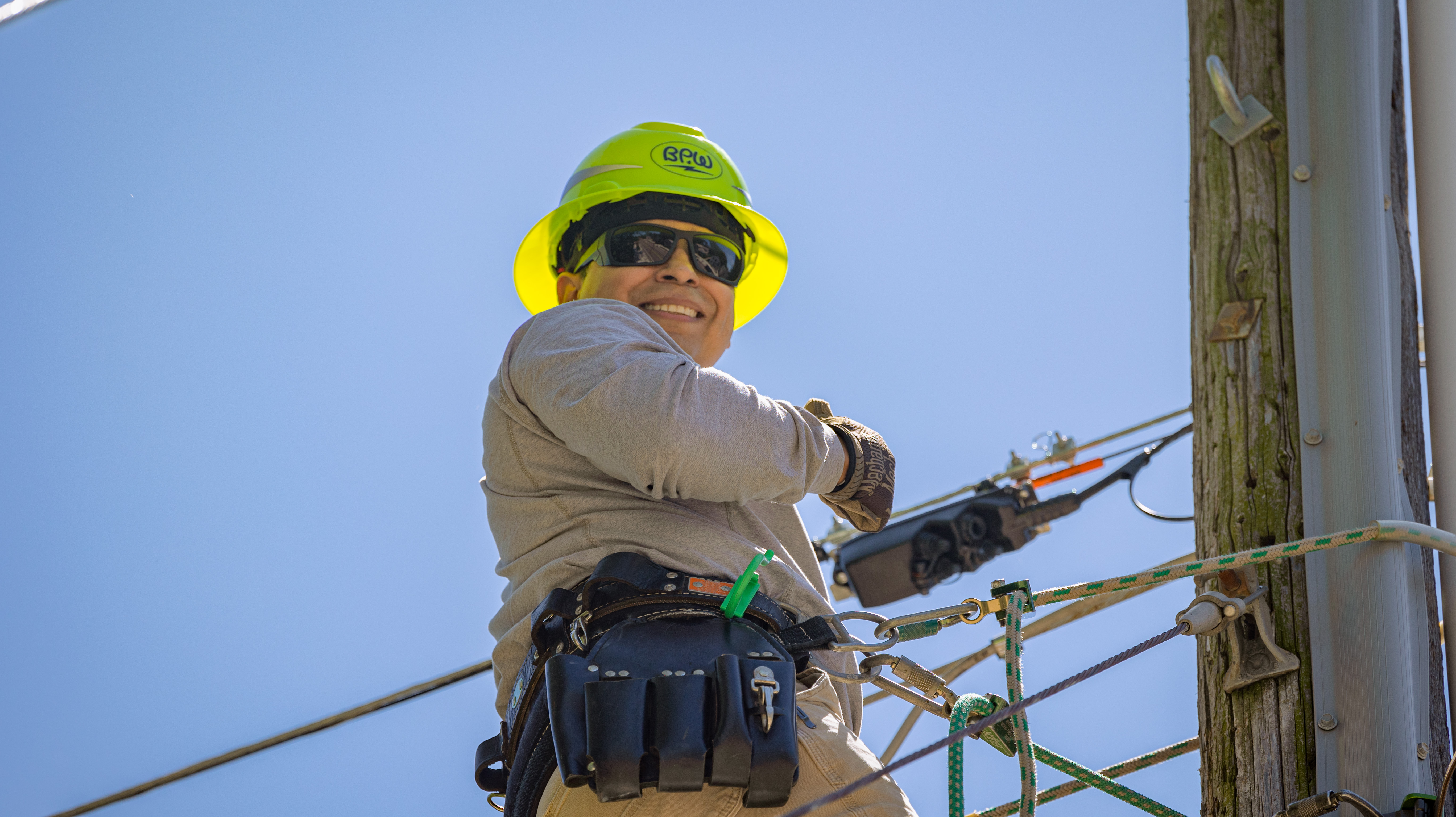 A fiber optic worker wears a hard hat and gloves. He smiles down from a utility pole, which he is harnessed to.